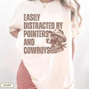 May include: Ivory-colored t-shirt with the text "EASILY DISTRACTED BY POINTERS AND COWBOYS" in brown. A brown dog wearing a cowboy hat and bandana is featured on the shirt. The shirt is a casual style.