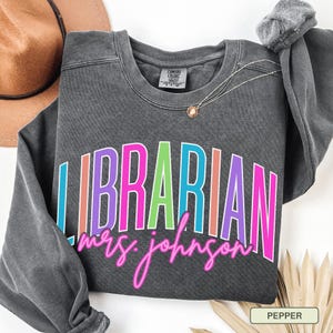 May include: A dark gray sweatshirt with the word "LIBRARIAN" in colorful block letters and "mrs. johnson" in pink script. A gold necklace and a brown hat are visible. The sweatshirt is labeled "Pepper".
