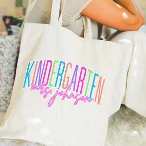 May include: Cream-colored tote bag with the words "KINDERGARTEN mrs. johnson" in colorful, arched text. The bag has long handles and a rectangular "TOTE BAG" label at the bottom. The bag is held by a person wearing a neutral-colored top and white pants.