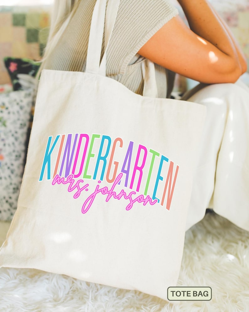 May include: Cream-colored tote bag with the words "KINDERGARTEN mrs. johnson" in colorful, arched text. The bag has long handles and a rectangular "TOTE BAG" label at the bottom. The bag is held by a person wearing a neutral-colored top and white pants.