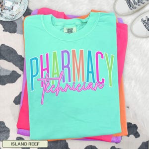 May include: A stack of colorful t-shirts, with a teal shirt on top. The teal shirt has the words "PHARMACY Technician" in a rainbow font. The other shirts are pink and orange. The shirt is labeled "ISLAND REEF".