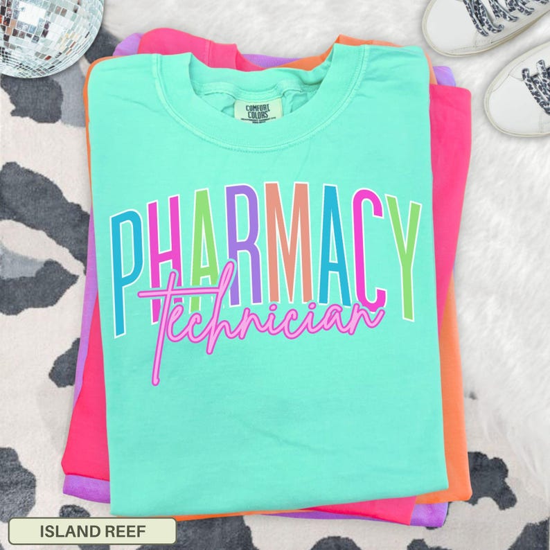 May include: A stack of colorful t-shirts, with a teal shirt on top. The teal shirt has the words "PHARMACY Technician" in a rainbow font. The other shirts are pink and orange. The shirt is labeled "ISLAND REEF".