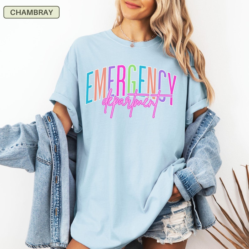 May include: Pale blue t-shirt featuring the text "EMERGENCY department" in a layered, multi-coloured design. The shirt has a round neck and short sleeves. A denim jacket is worn over the shoulders.
