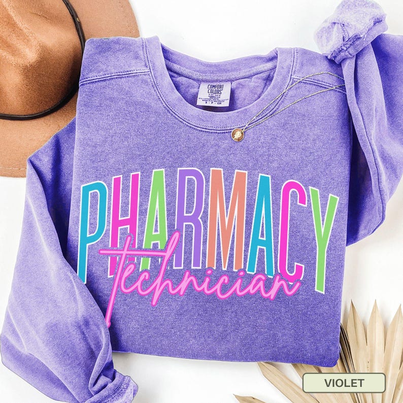 May include: A violet-colored sweatshirt with the words "PHARMACY Technician" in a colorful, outlined font. The sweatshirt has a crew neck and long sleeves. A gold necklace is draped on the front. A brown hat is visible in the background.