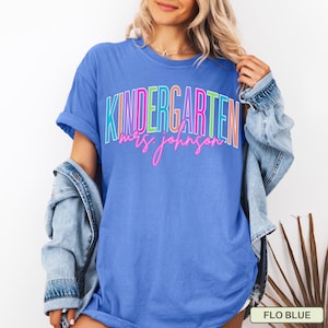 May include: A blue t-shirt with the word "KINDERGARTEN" in colorful block letters and "mrs. johnson" in pink script. The shirt is a solid blue color and has short sleeves. A denim jacket is draped over the shoulders.