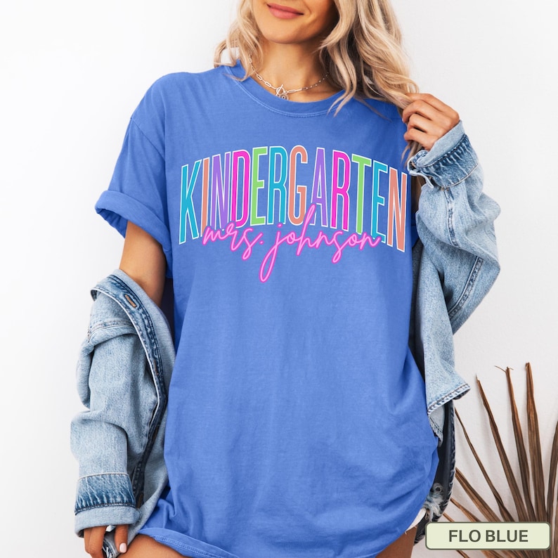 May include: A blue t-shirt with the word "KINDERGARTEN" in colorful block letters and "mrs. johnson" in pink script. The shirt is a solid blue color and has short sleeves. A denim jacket is draped over the shoulders.