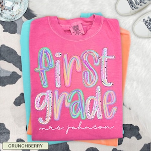 May include: A stack of t-shirts in shades of pink, orange, and turquoise. The top shirt is pink and features the words "First Grade" in a colorful, glittery font, with "mrs. johnson" below. The shirt is labeled "CRUNCHBERRY".