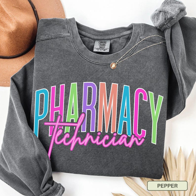 May include: A dark gray sweatshirt with the words "PHARMACY Technician" in a colorful, outlined font. The sweatshirt is a Comfort Colors brand and has a crew neck. A gold necklace with a pendant is draped on the sweatshirt.