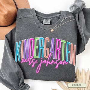 May include: A dark gray sweatshirt with the word "KINDERGARTEN" in colorful block letters and "mrs. johnson" in pink script. A gold necklace and a brown hat are also visible.