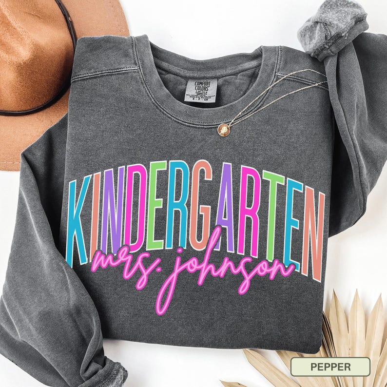 May include: A dark gray sweatshirt with the word "KINDERGARTEN" in colorful block letters and "mrs. johnson" in pink script. A gold necklace and a brown hat are also visible.