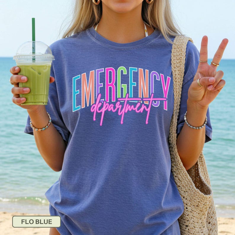 May include: A blue t-shirt featuring the text "EMERGENCY department" in a vibrant, neon-style font. The shirt is plain and has short sleeves. The background shows a beach and the sea, suggesting an outdoor setting.