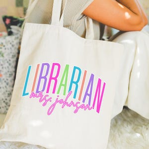 May include: Cream-colored tote bag with the word "LIBRARIAN" in colorful, arched letters. Below, in a cursive font, is "mrs. johnson". The bag has two handles and a small tag that says "TOTE BAG".