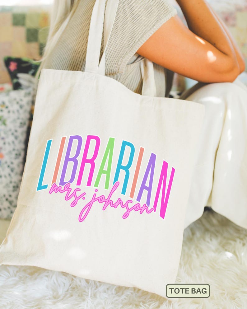 May include: Cream-colored tote bag with the word "LIBRARIAN" in colorful, arched letters. Below, in a cursive font, is "mrs. johnson". The bag has two handles and a small tag that says "TOTE BAG".