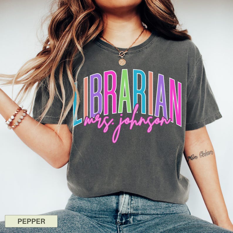 May include: A dark gray t-shirt with the word "LIBRARIAN" in colorful block letters and "mrs. johnson" in neon pink script. The shirt is a casual style, suitable for everyday wear.