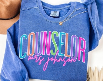 Könnte beinhalten: Ein blaues Sweatshirt mit Rundhalsausschnitt, das das Wort COUNSELOR in großen, bunten Blockbuchstaben und den Text mrs. johnson in einer leuchtend pinken Schreibschrift zeigt. Das Kleidungsstück hat ein Comfort Colors Etikett und eine Goldkette.