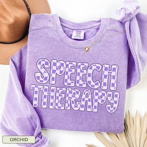 May include: A lavender crewneck sweatshirt with a washed-out finish featuring the text SPEECH THERAPY in a purple and white checkered pattern. The garment has a Comfort Colors brand label at the inner neckline and long sleeves.