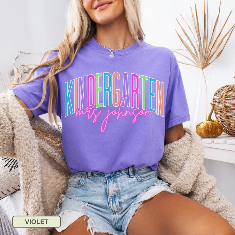 May include: A violet-colored t-shirt with the word "KINDERGARTEN" in a colorful, outlined font. Below the word is the name "Mrs. Johnson" in a cursive, pink font. The shirt is a short-sleeved tee.