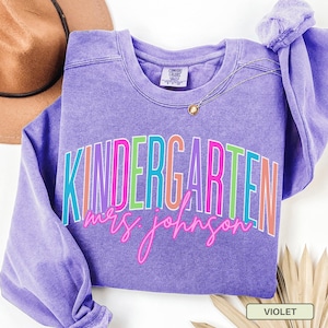 May include: A violet-colored sweatshirt with the word "KINDERGARTEN" in a colorful, layered font. Below the word is the name "mrs. johnson" in a cursive font. A gold necklace and a brown hat are visible in the background.