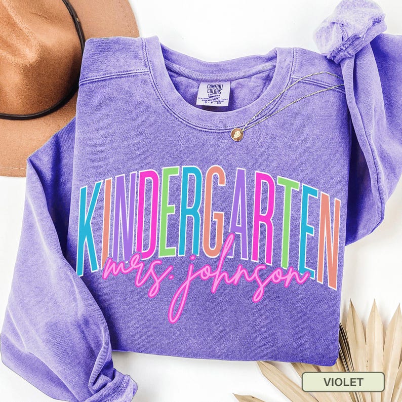 May include: A violet-colored sweatshirt with the word "KINDERGARTEN" in a colorful, layered font. Below the word is the name "mrs. johnson" in a cursive font. A gold necklace and a brown hat are visible in the background.