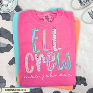 May include: A bright pink short-sleeved t-shirt featuring the text "ELL CREW" in large, multi-colored, marbled letters with black speckles. Below the main text, the words "mrs. johnson" are printed in a white, cursive script font.