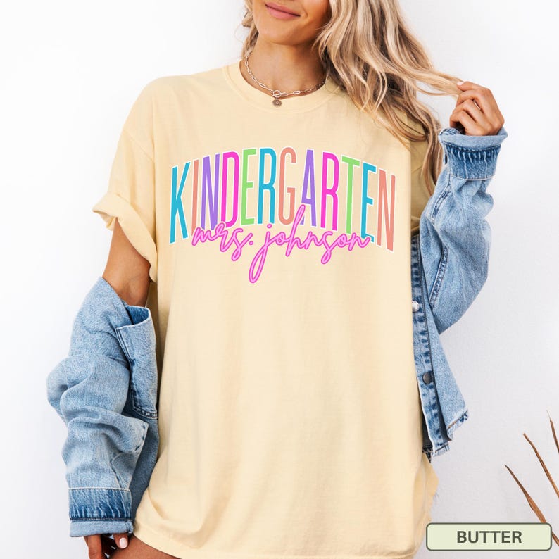 May include: A light yellow t-shirt with the word "KINDERGARTEN" in colorful block letters and "mrs. johnson" in cursive. A denim jacket is draped over the shoulders. The shirt is a butter color.