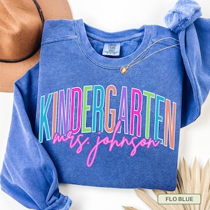 May include: A blue sweatshirt with the word "KINDERGARTEN" in colorful block letters and "mrs. johnson" in pink cursive. A gold necklace and a brown hat are visible.