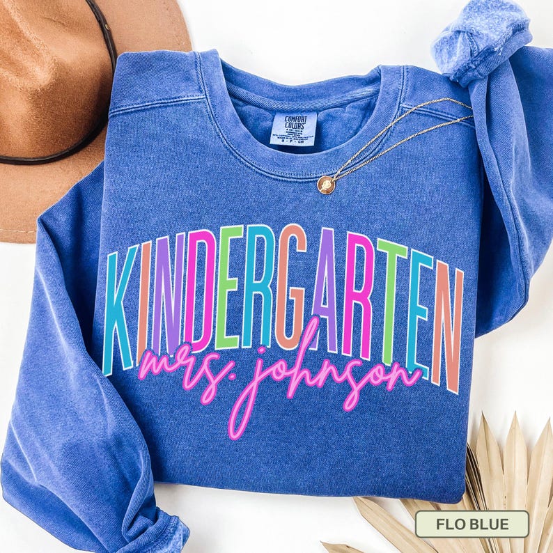 May include: A blue sweatshirt with the word "KINDERGARTEN" in colorful block letters and "mrs. johnson" in pink cursive. A gold necklace and a brown hat are visible.