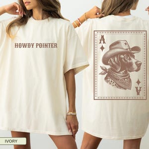 May include: Ivory-colored t-shirt with the text "HOWDY POINTER" in brown. The back features a brown illustration of a dog wearing a cowboy hat and bandana, framed by a playing card design with the letter "A".