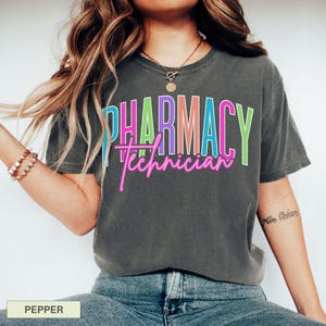 May include: A dark gray t-shirt with the words "PHARMACY Technician" in a colorful, layered design. The word "PHARMACY" is in a block letter style with blue, pink, purple, and green colors. The word "Technician" is in a neon pink script.