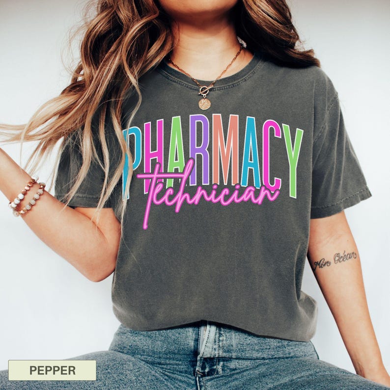 May include: A dark gray t-shirt with the words "PHARMACY Technician" in a colorful, layered design. The word "PHARMACY" is in a block letter style with blue, pink, purple, and green colors. The word "Technician" is in a neon pink script.