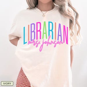 May include: Ivory-colored t-shirt with the word "LIBRARIAN" in colorful block letters and "mrs. johnson" in a cursive font. The shirt is a casual style, suitable for everyday wear.