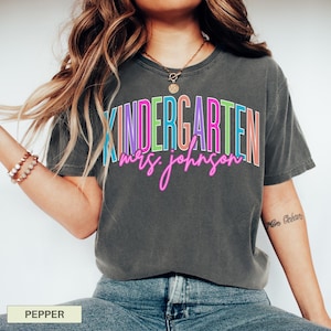 May include: A dark gray t-shirt with the word "KINDERGARTEN" in colorful block letters and "mrs. johnson" in pink cursive. The shirt is a comfortable, casual style.