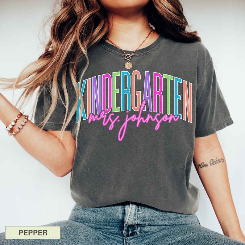 May include: A dark gray t-shirt with the word "KINDERGARTEN" in colorful block letters and "mrs. johnson" in pink cursive. The shirt is a comfortable, casual style.