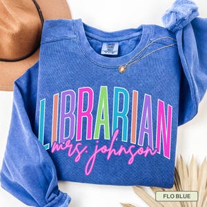 May include: A blue sweatshirt with the word "LIBRARIAN" in colorful block letters. Below the word "LIBRARIAN" is the name "mrs. johnson" in a cursive font. A gold necklace and a brown hat are also visible.