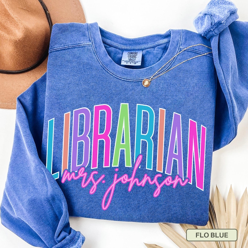 May include: A blue sweatshirt with the word "LIBRARIAN" in colorful block letters. Below the word "LIBRARIAN" is the name "mrs. johnson" in a cursive font. A gold necklace and a brown hat are also visible.