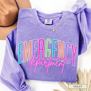 May include: A violet sweatshirt featuring the text "EMERGENCY department" in a multi-coloured, outlined font. The sweatshirt has a crew neck and long sleeves. A gold necklace is draped on the front. A brown hat is visible in the background.