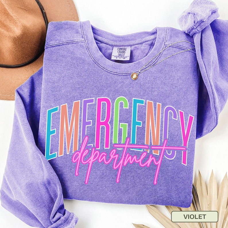 May include: A violet sweatshirt featuring the text "EMERGENCY department" in a multi-coloured, outlined font. The sweatshirt has a crew neck and long sleeves. A gold necklace is draped on the front. A brown hat is visible in the background.