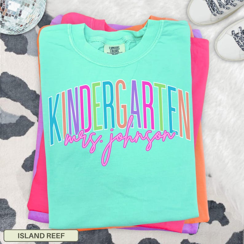 May include: A turquoise Comfort Colors t-shirt with the word "KINDERGARTEN" in colorful block letters and "mrs. johnson" in pink script. The shirt is folded on top of other brightly colored shirts. The bottom of the shirt reads "ISLAND REEF".