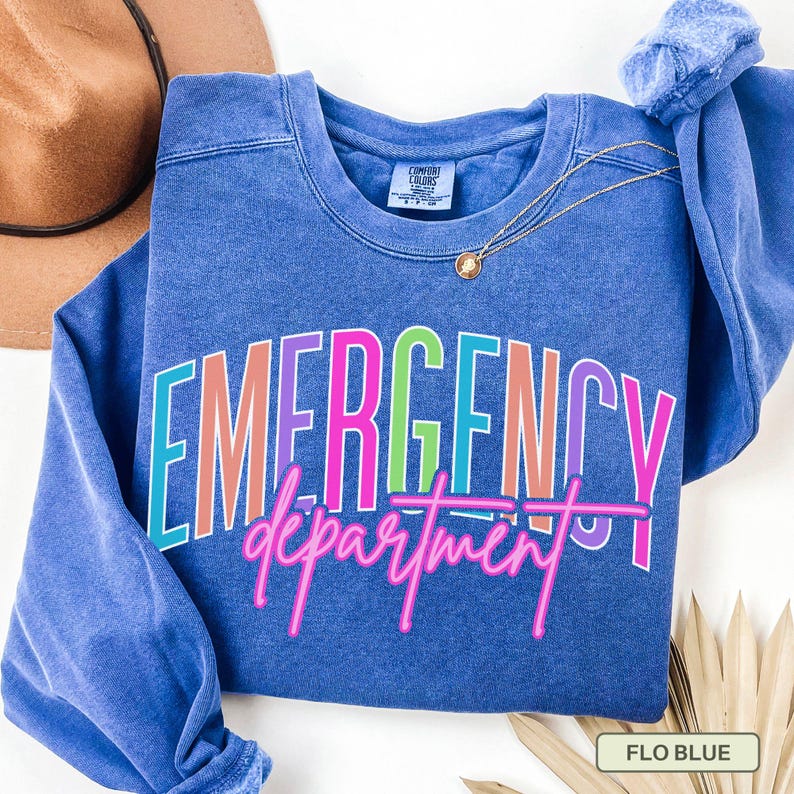 May include: A blue sweatshirt with the words "EMERGENCY department" in colourful text. The sweatshirt has a crew neck and long sleeves. A gold necklace and a brown hat are also visible.