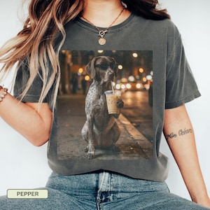 May include: Dark grey t-shirt featuring a graphic of a dog wearing sunglasses and holding an iced coffee. The dog is brown and white, set against a blurred city background. The shirt is labeled "PEPPER" at the bottom.