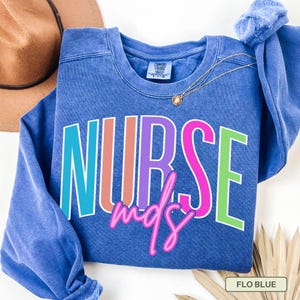 May include: A blue crewneck sweatshirt with the word "NURSE" in colorful block letters and "mds" in pink script. The sweatshirt has a vintage wash and a Comfort Colors tag. A gold necklace and a brown hat are also visible.