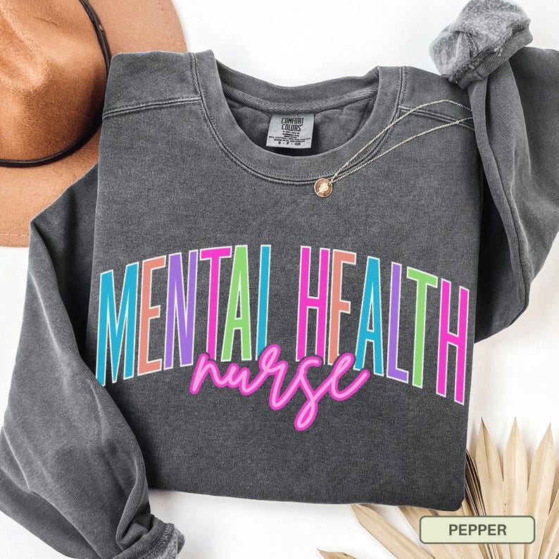 May include: A dark gray sweatshirt with the words "MENTAL HEALTH nurse" in a colorful, outlined font. The sweatshirt has a crew neck and long sleeves. A gold necklace with a pendant is draped on the shirt.