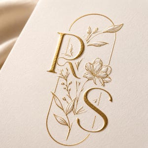 May include: A cream-coloured card featuring a gold foil embossed design of the letters R and S. The letters are intertwined with delicate, raised white floral and botanical line art, all enclosed within a thin, gold-lined arch shape.