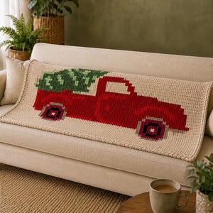 May include: A rectangular crochet blanket in a light beige color featuring a pixelated graphic of a red pickup truck carrying a green pine tree. The blanket has a textured stitch pattern and a finished border, displaying a festive holiday theme.