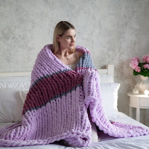 Könnte beinhalten: Eine weiche, dicke Strickdecke in Lavendel-, Rosa- und Grautönen. Die Decke liegt über einem Bett und eine Person sitzt auf dem Bett.