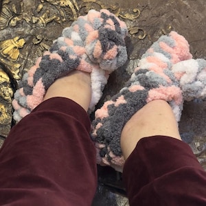 May include: A pair of cozy, handmade slippers in a mix of pink, gray, and white yarn. The slippers appear soft and plush, perfect for lounging. The chunky knit design adds texture and warmth to these comfortable house shoes.