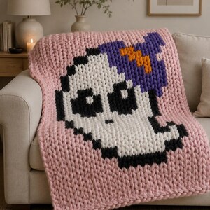 May include: A chunky knit blanket in light pink featuring a large pixelated skull design in white and black. The skull is topped with a purple and orange pixelated hat. The blanket is crafted from thick yarn with a visible, textured stitch pattern.