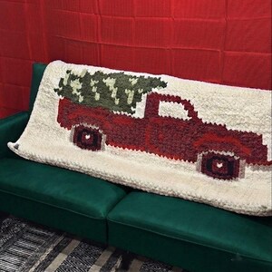 Peut inclure: Un plaid rectangulaire de couleur crème présentant un grand motif texturé de style pixelisé représentant un pick-up rouge transportant un sapin de Noël vert foncé dans sa benne. La couverture a une texture épaisse, touffetée ou shaggy.