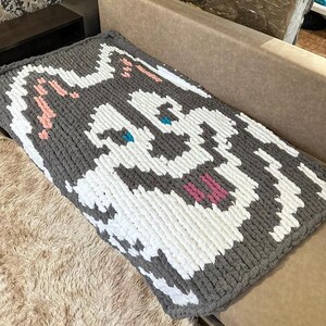 May include: A gray and white knitted throw blanket with a cartoon dog face design. The dog's eyes are blue, and the nose and mouth are pink.