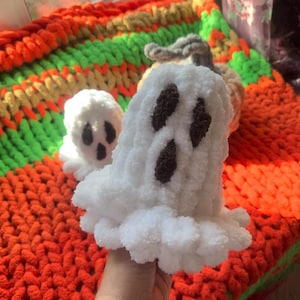 May include: Two white crocheted ghost ornaments with brown eyes, sitting on a colorful knitted blanket.